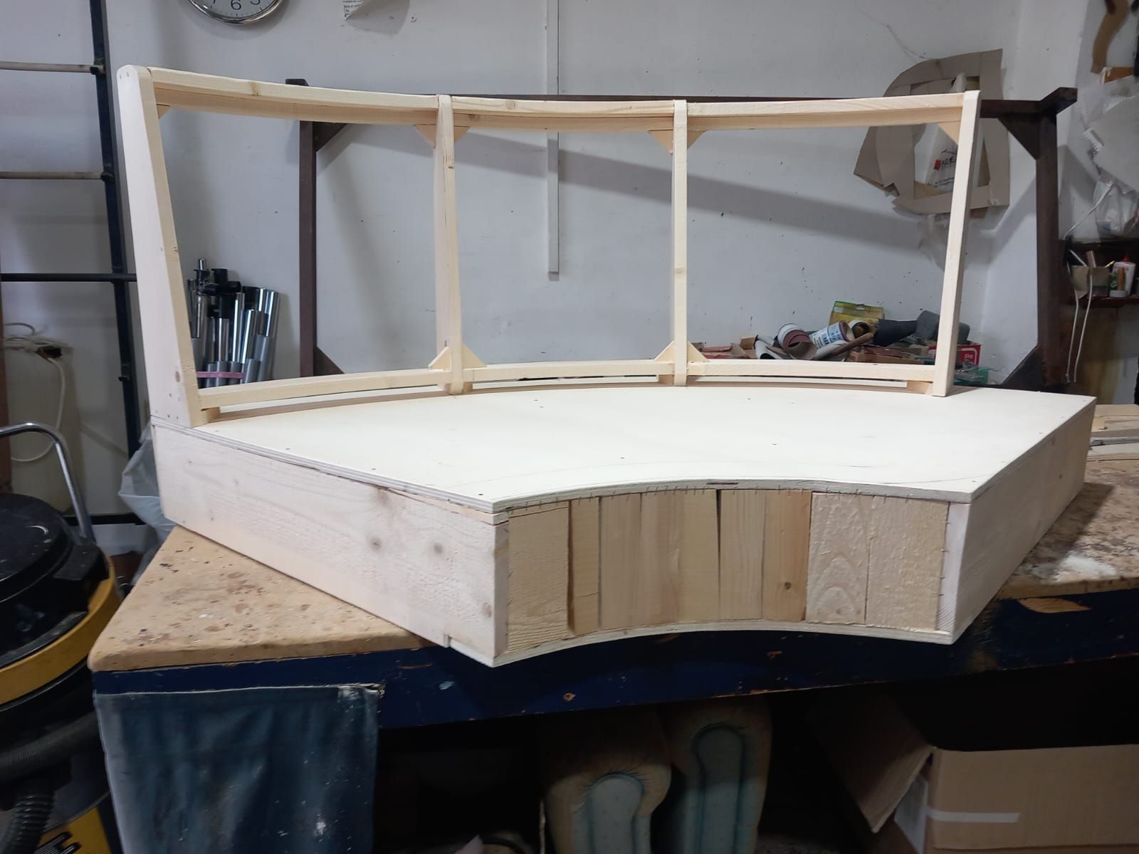 Un divano componibile in legno parzialmente costruito in un laboratorio. La base curva è in legno chiaro e la struttura dello schienale è in fase di costruzione.