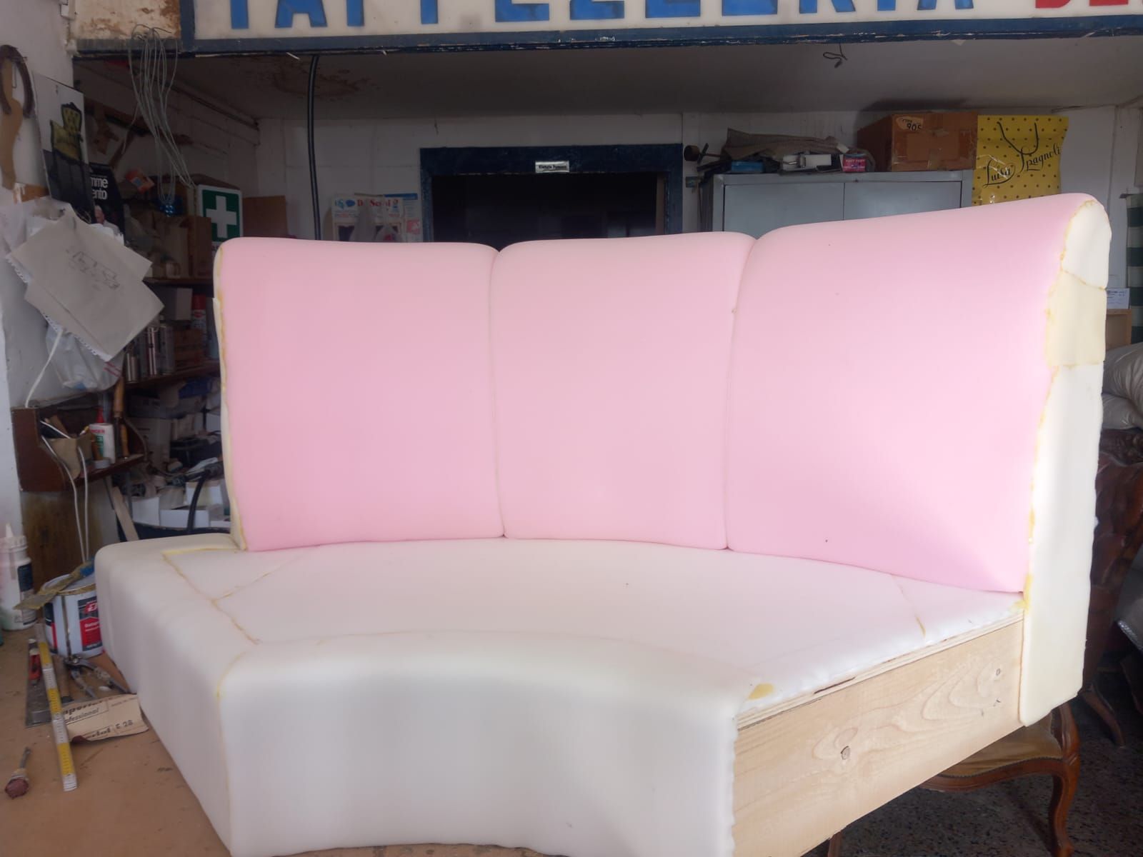 Un divano parzialmente imbottito rosa e bianco in un ambiente da laboratorio. Presenta schienale e seduta curvi, con struttura in legno a vista.
