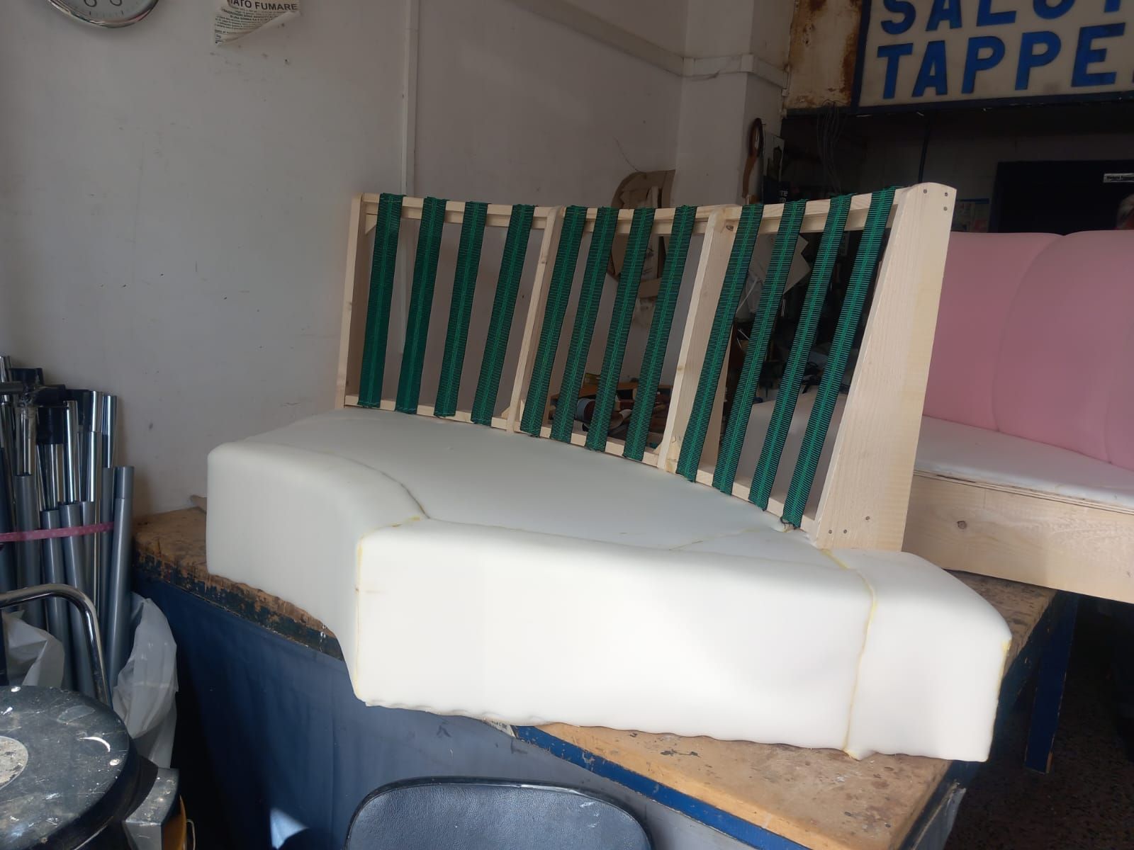 Un divano in schiuma bianca parzialmente costruito, con struttura in legno e cinghie verdi. Si trova in un laboratorio di tappezzeria.
