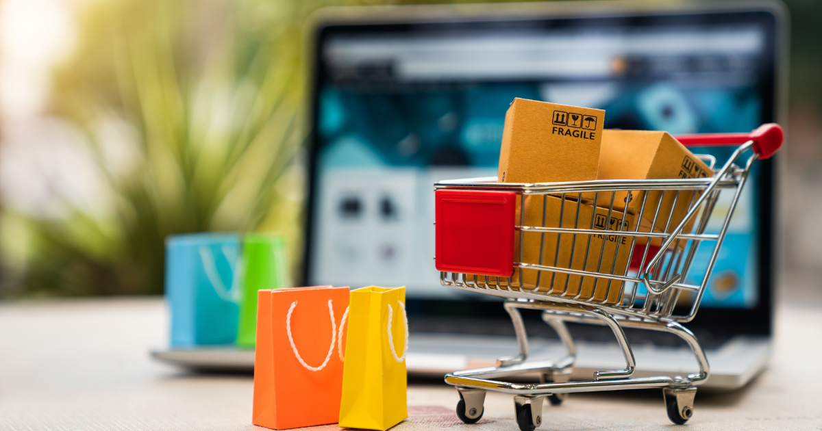 Mini shopping cart with parcels and colorful bags in front of a laptop displaying an e-commerce site.