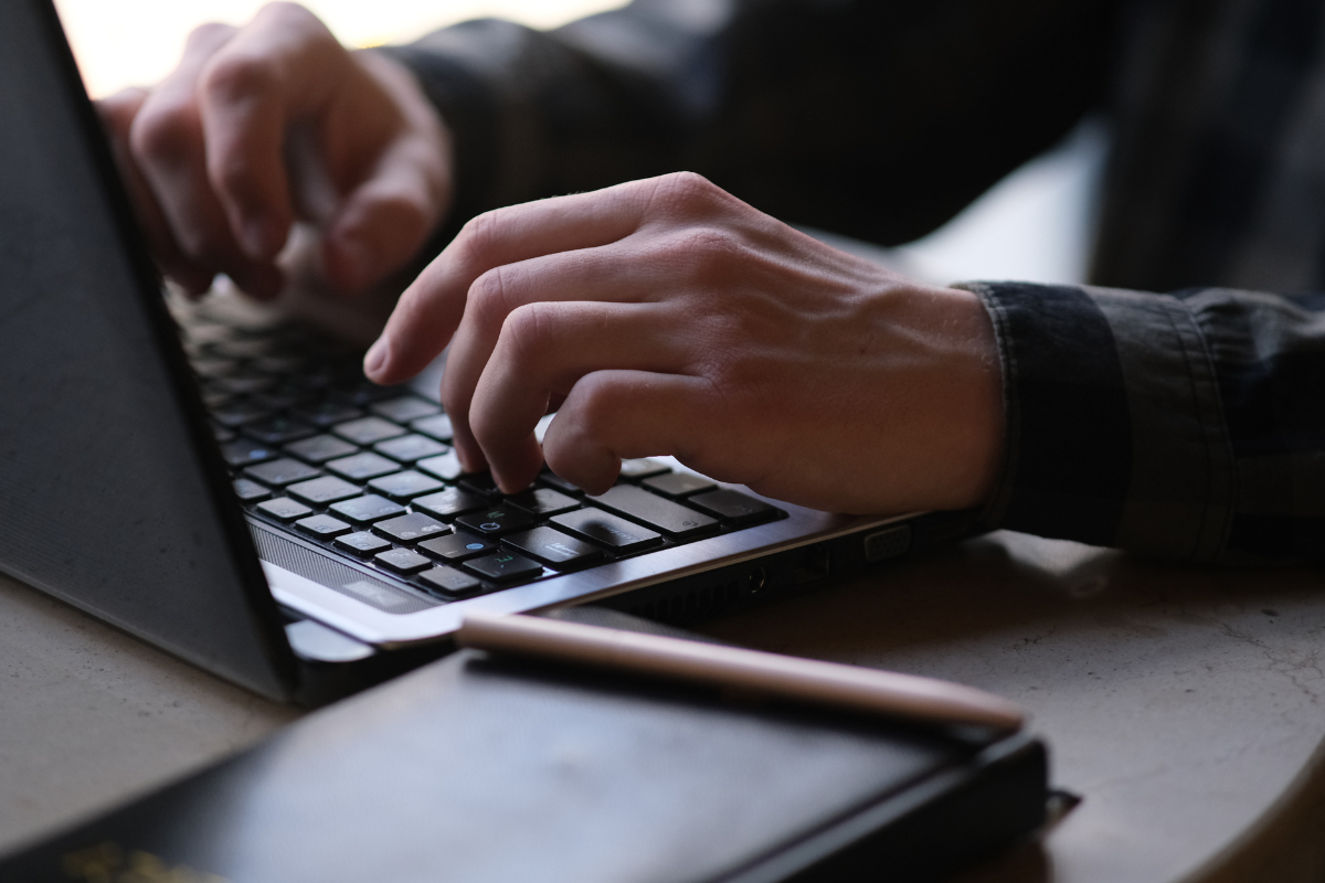 A close-up of hands typing on a laptop keyboard, with a notebook and pen on the table nearby.
