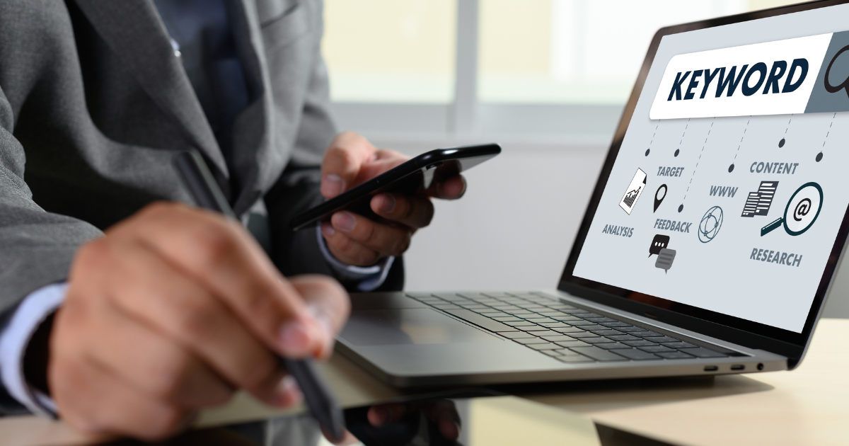 Person using phone and stylus while viewing keyword strategy on a laptop screen for SEO and content research.