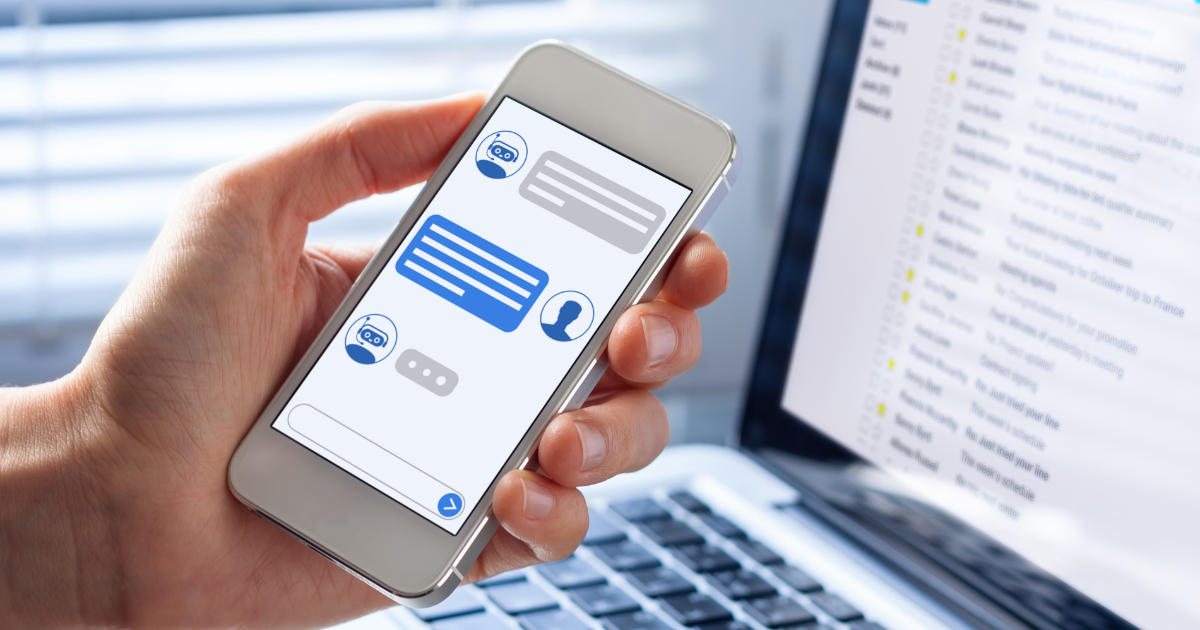 A person holds a smartphone displaying a chatbot conversation, with a laptop showing an open email inbox in the background.