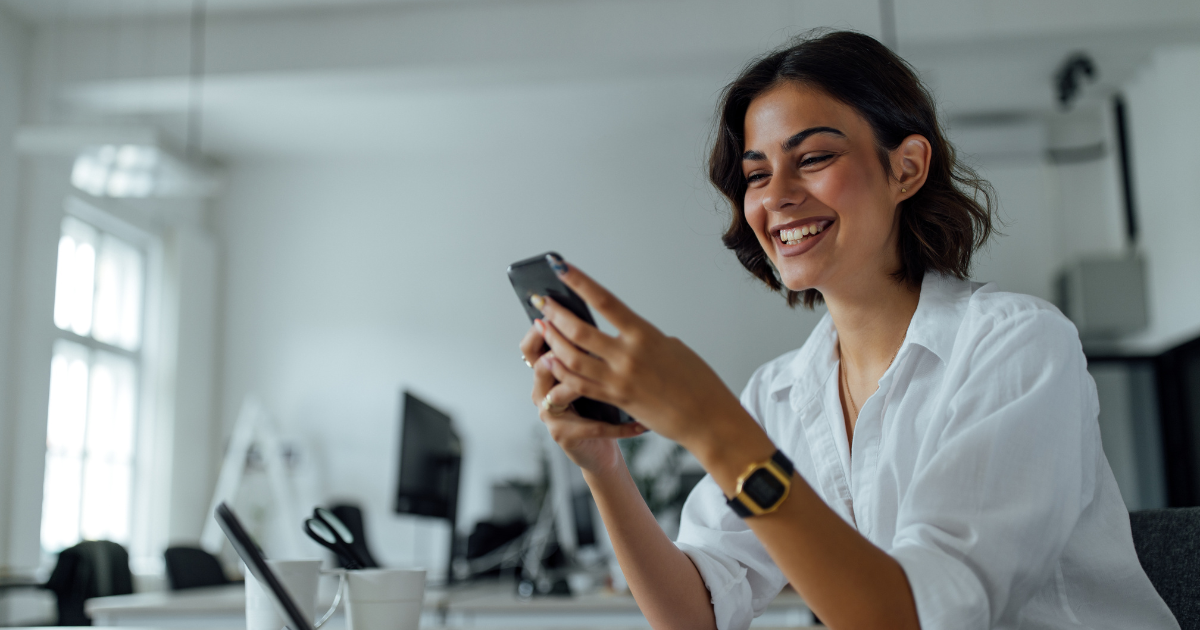 Smiling woman using smartphone at desk, reflecting strategic buying behavior in a work setting.