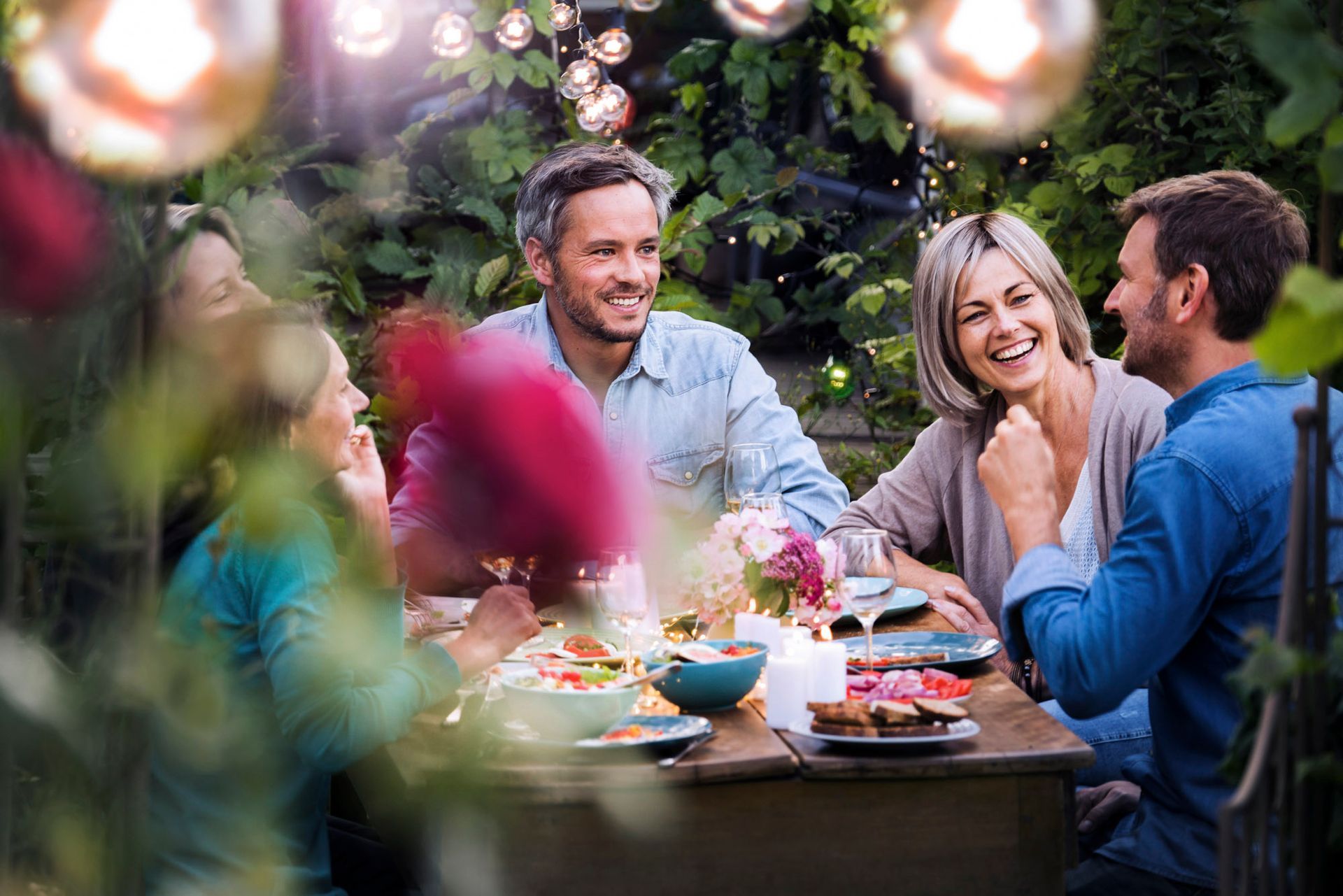 Friends laughing and eating at a table outdoors, string lights overhead, flowers and food present.