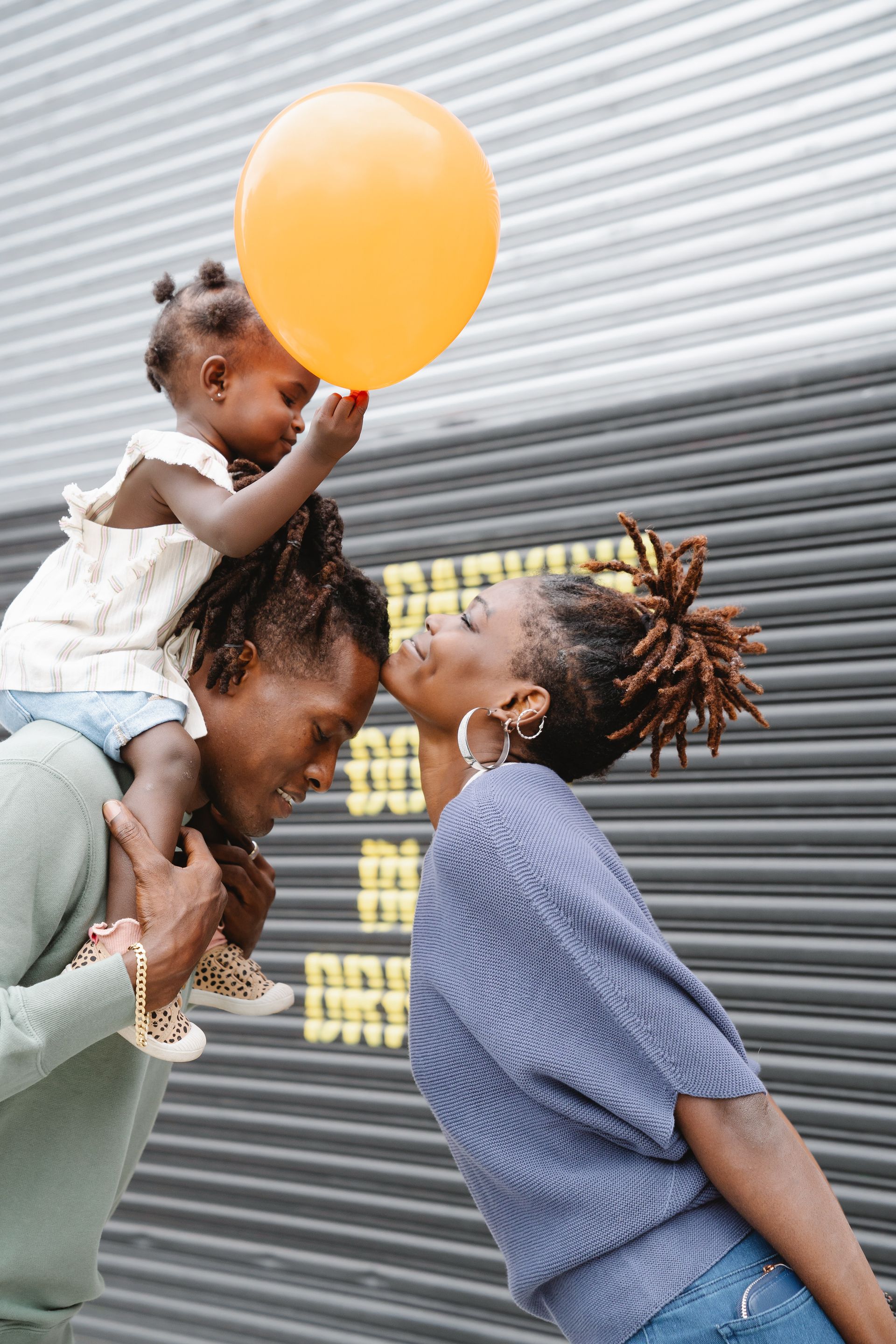 Adult lifting a smiling child with a yellow balloon against a corrugated metal wall