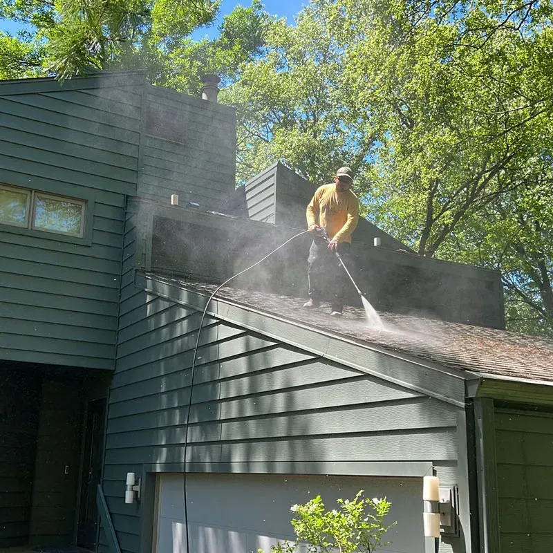 A person in yellow sprays a roof, releasing mist. The house has green siding, trees surround it, and a clear sky.