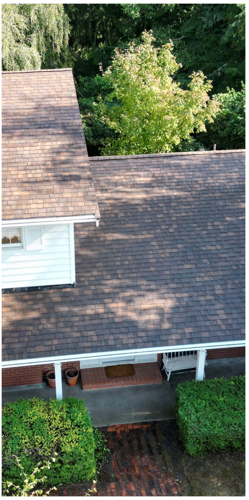 Overhead view of a house with a brown roof and a white porch with green bushes and trees in the background.