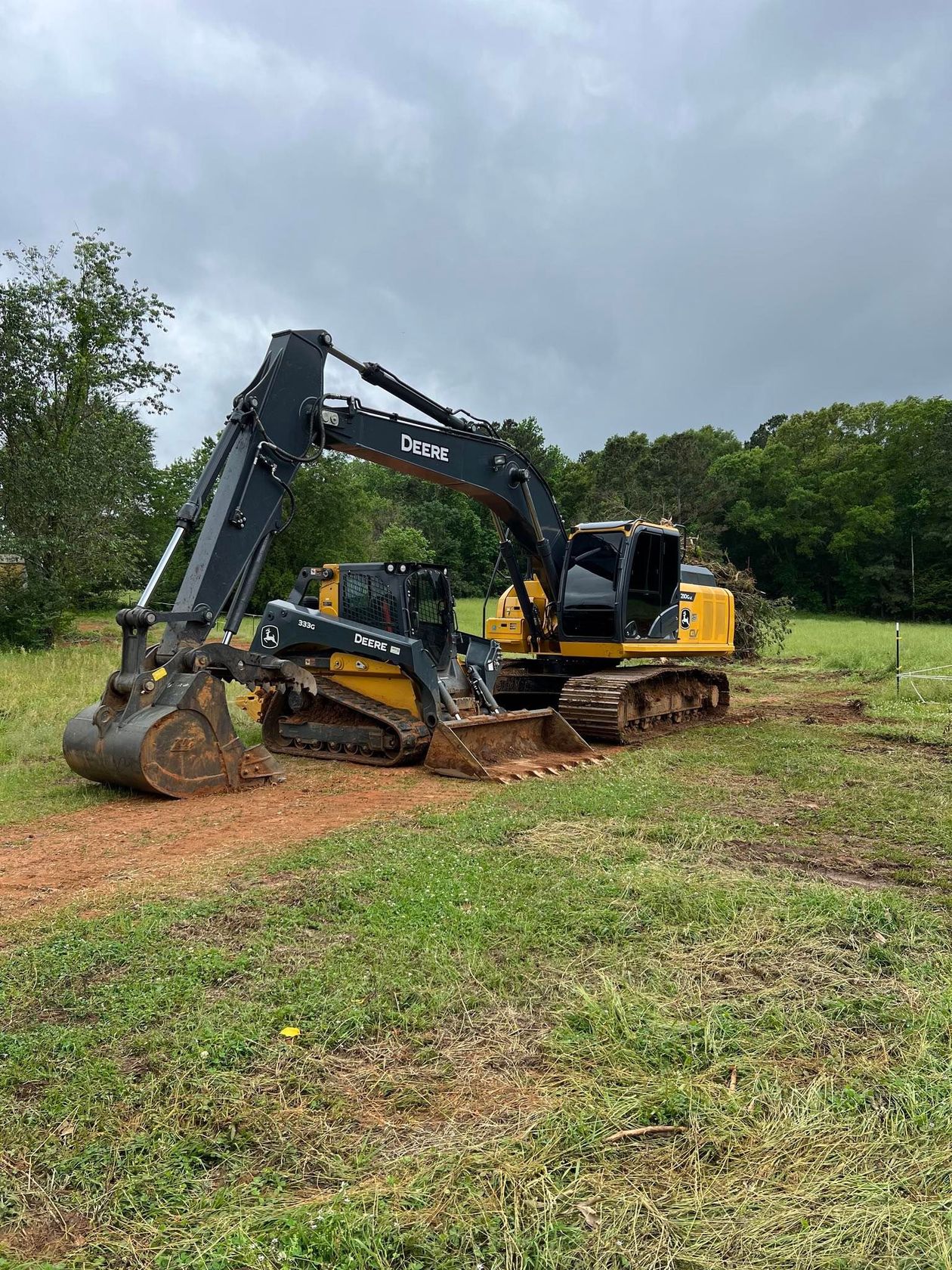 A large John Deere excavator digging in a grassy area, under an overcast sky.