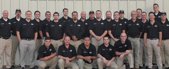 Group of men in black shirts and khaki pants pose in front of a wall. Some are kneeling.
