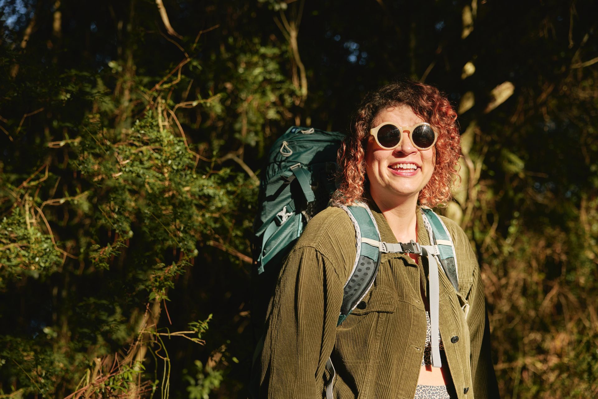 A woman wearing sunglasses and a backpack is standing in the woods.