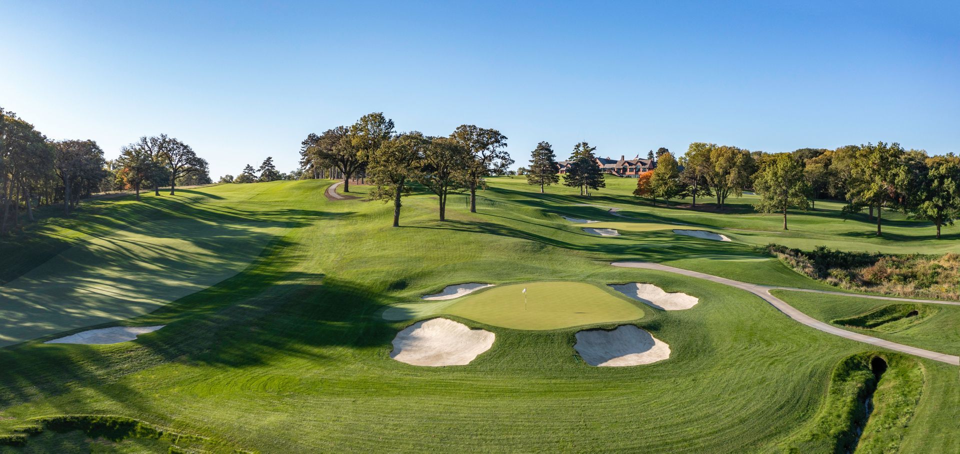 A golf course on a sunny day, with green grass, sand bunkers, and trees. Blue sky in the background.
