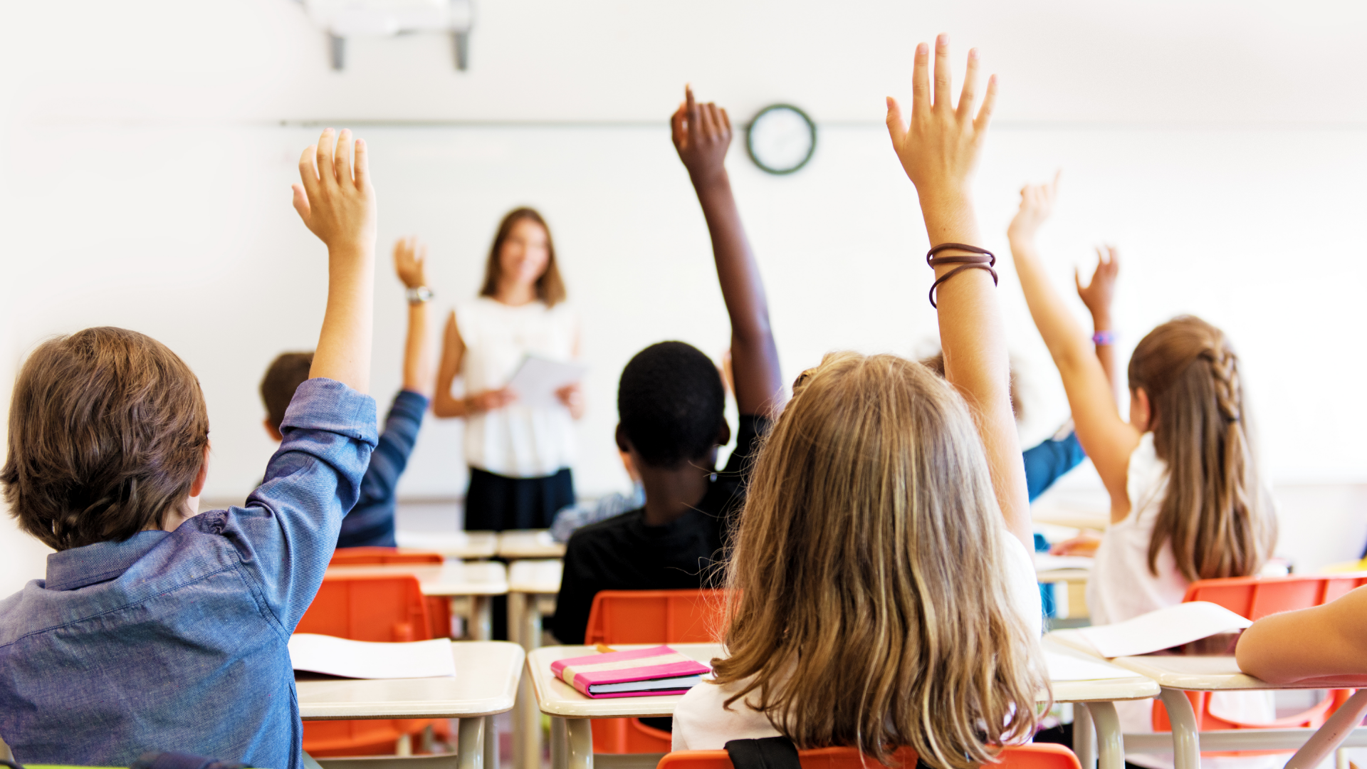 children at desks in classroom with arms raised toward blurred teacher