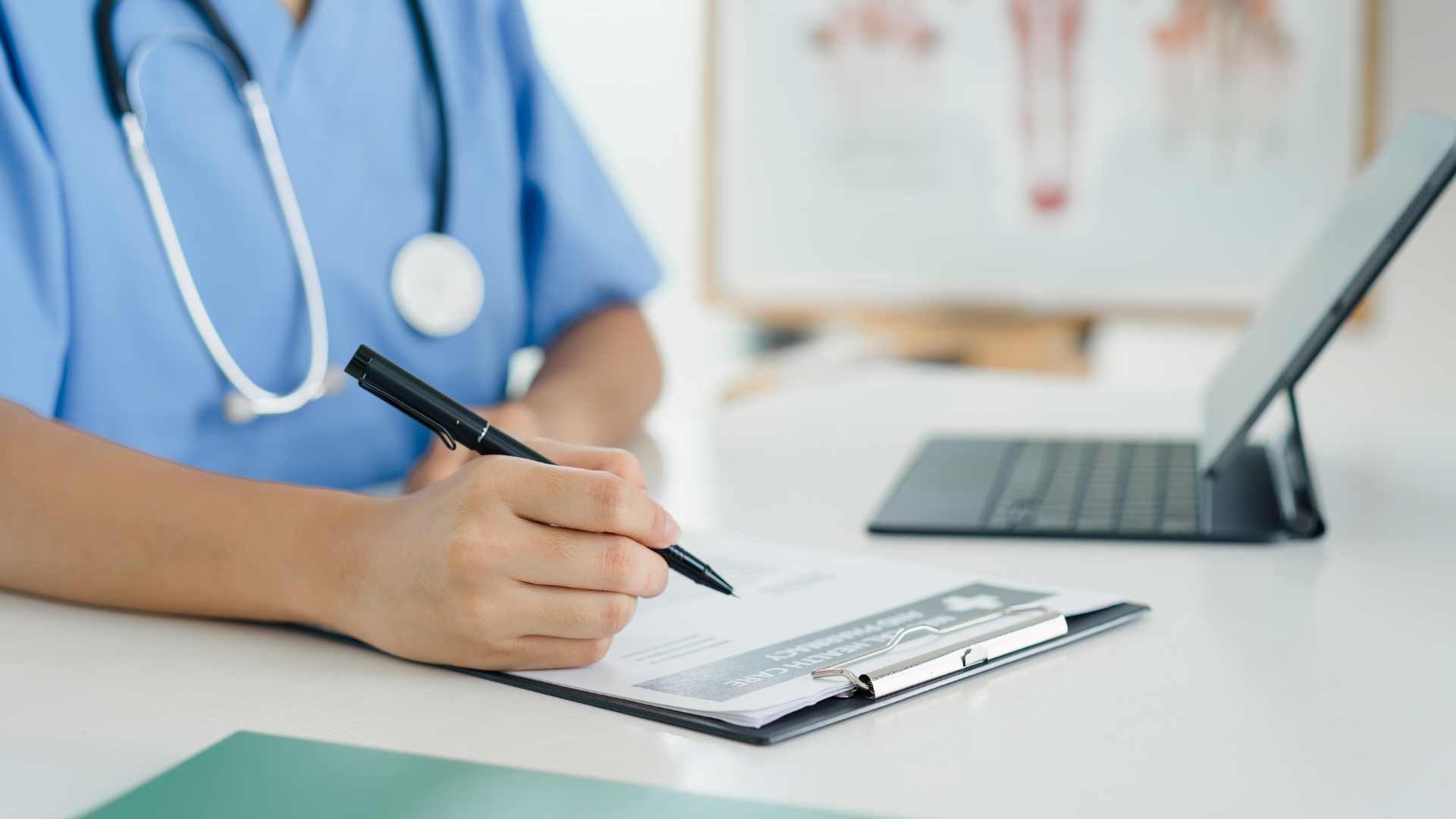 Medical professional in blue scrubs, writing on a clipboard, stethoscope around neck, tablet nearby.