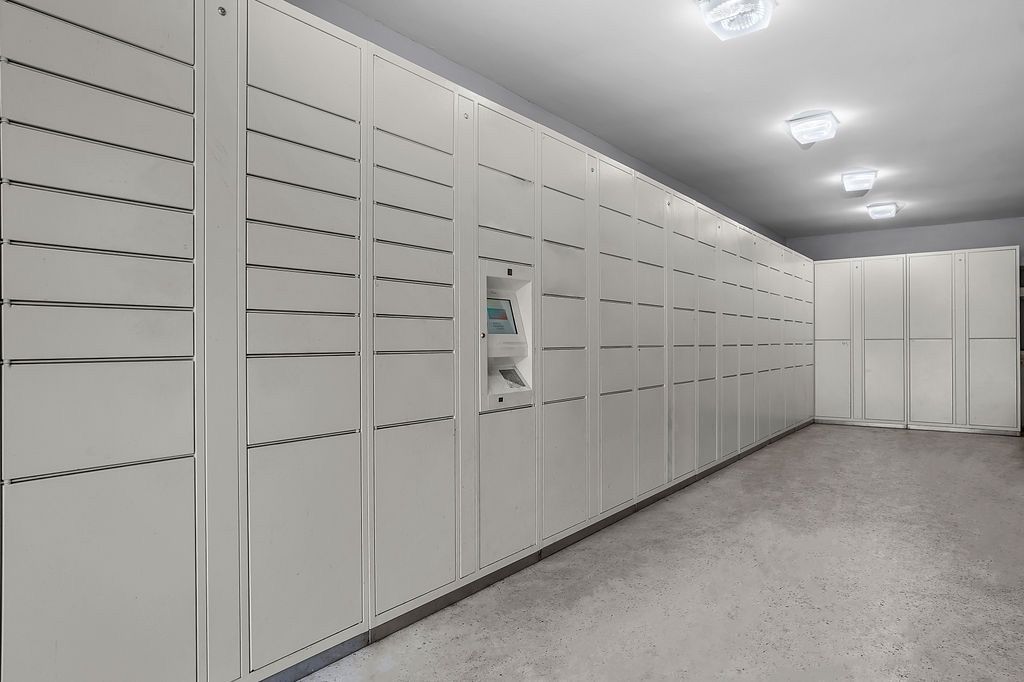 Long hallway of white storage lockers with a central package kiosk in a building mailroom.