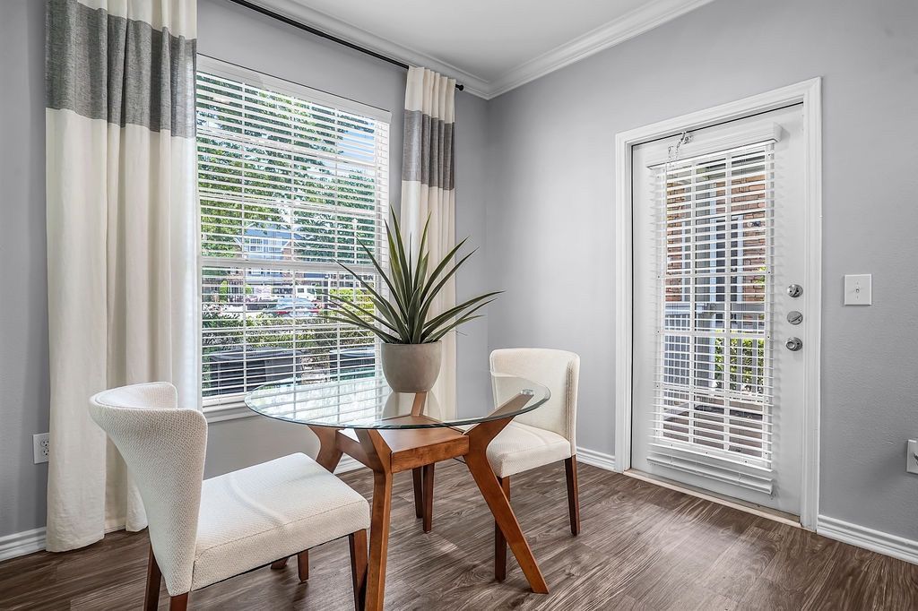 Interior dining area with a glass round table, two cream chairs, a plant, and large window with blinds.