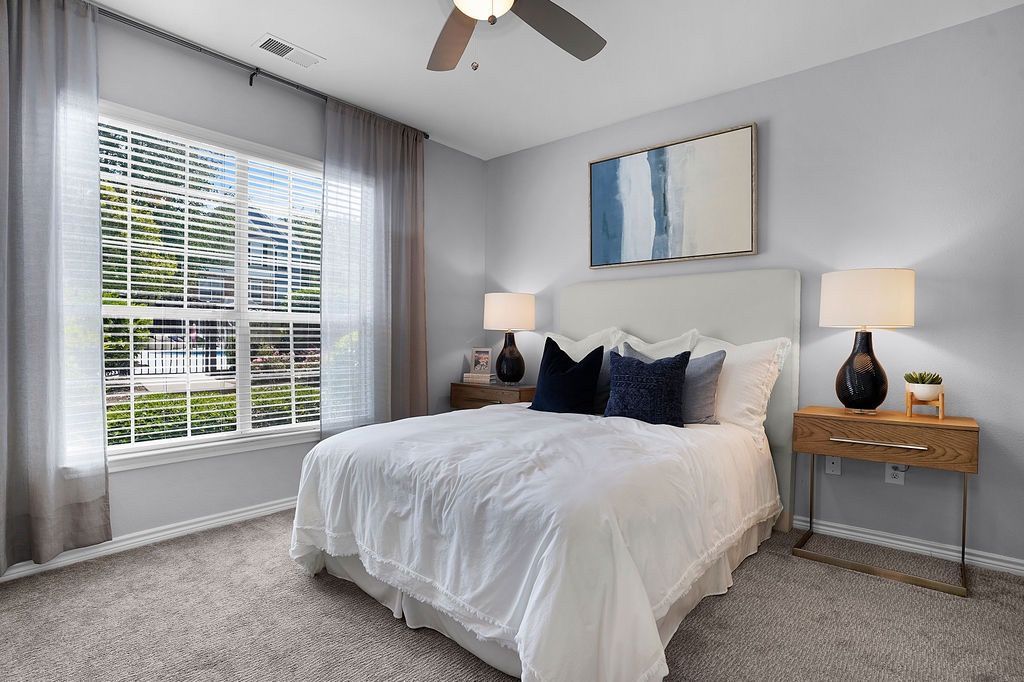 Bright bedroom with a white bed, two wooden nightstands, matching lamps, and a large window with sheer curtains.