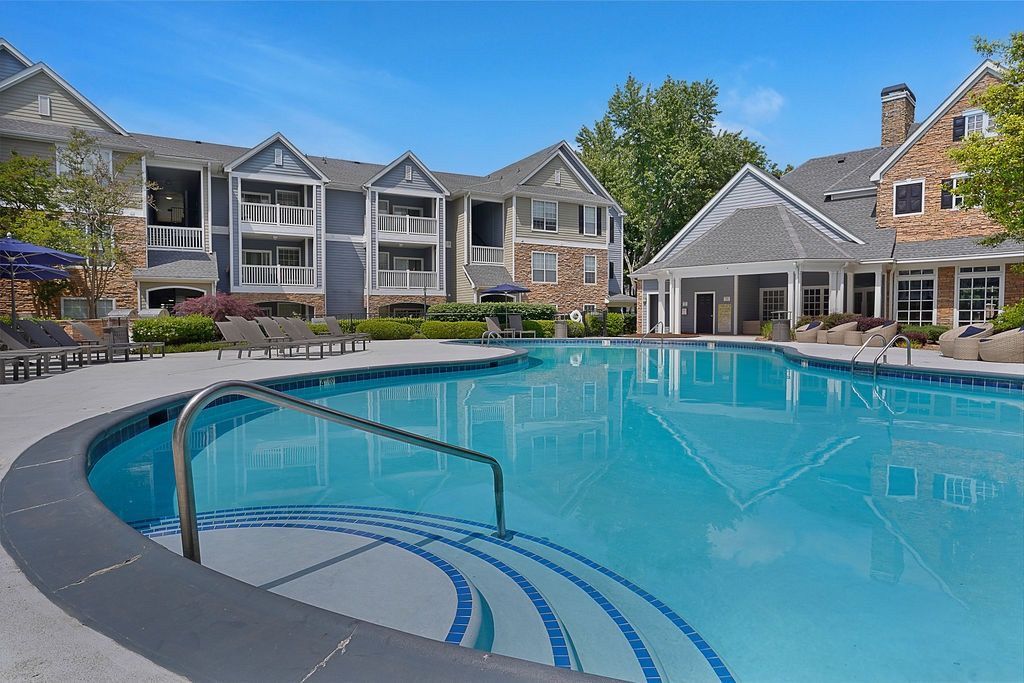 Outdoor community pool with curved steps and a metal handrail, surrounded by lounge chairs and apartment buildings.