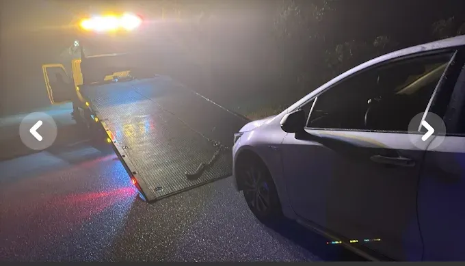 White car being loaded onto a tow truck at night, illuminated by the truck's flashing lights.