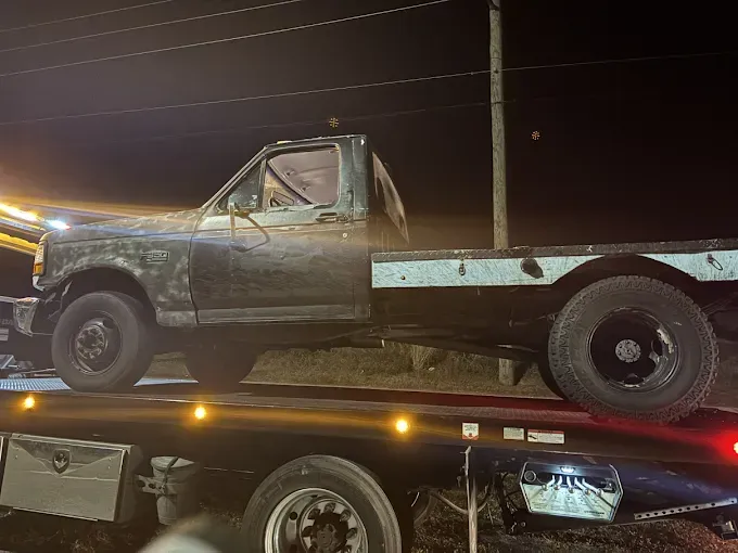 A weathered, dark pickup truck being towed on a flatbed tow truck at night.