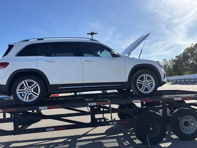 White SUV with open hood on a car hauler trailer, under a partly cloudy blue sky.