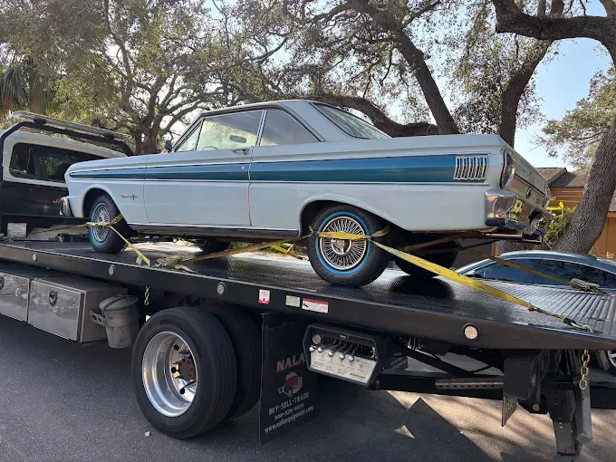 Classic light blue Ford Falcon on a flatbed tow truck. The car is secured with yellow straps. Daytime, outdoors.