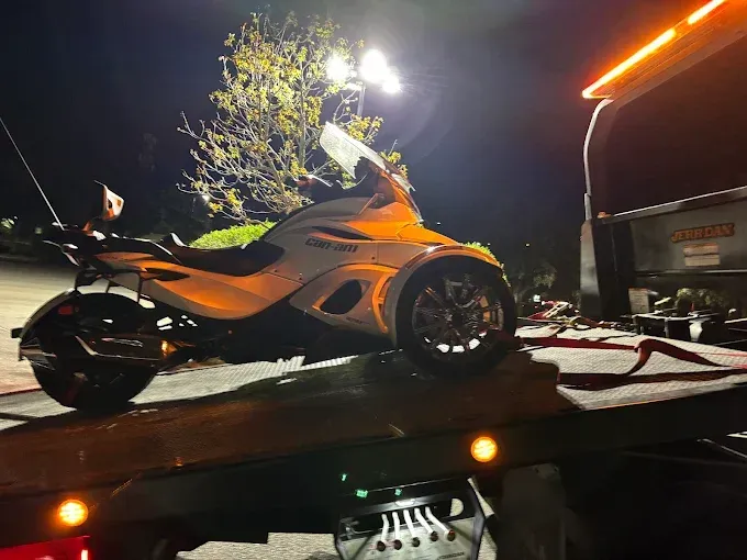 White three-wheeled motorcycle being towed on a flatbed truck at night.