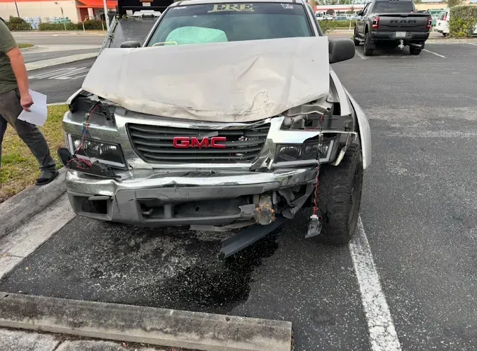 Damaged silver GMC truck after a collision, front end severely crushed, parked in a lot.