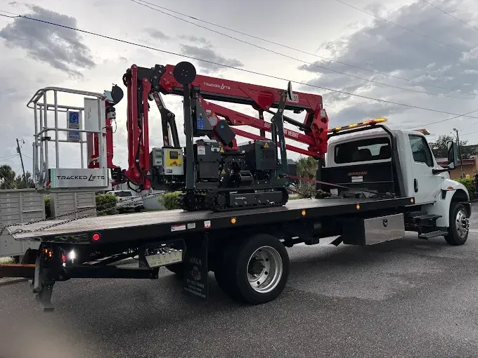 Flatbed truck transporting a mini excavator and aerial lift on a cloudy day.