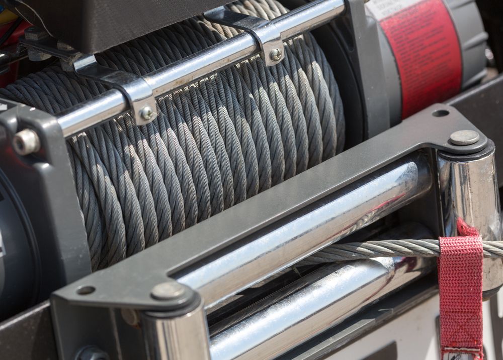 Close-up of a winch with coiled steel cable. Gray metal frame, red strap visible.