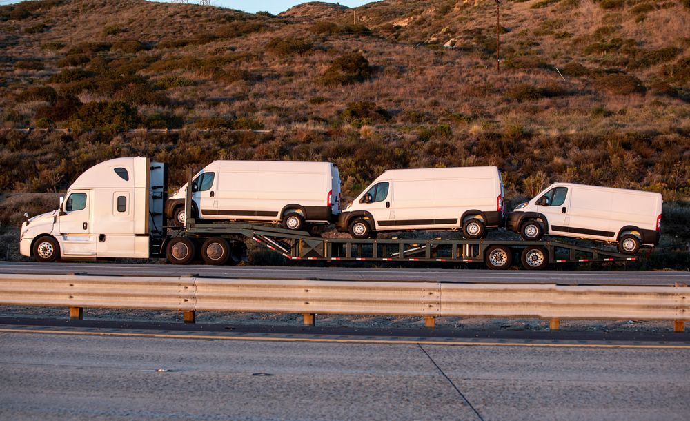 White semi-truck hauling three white vans on a flatbed trailer along a highway.