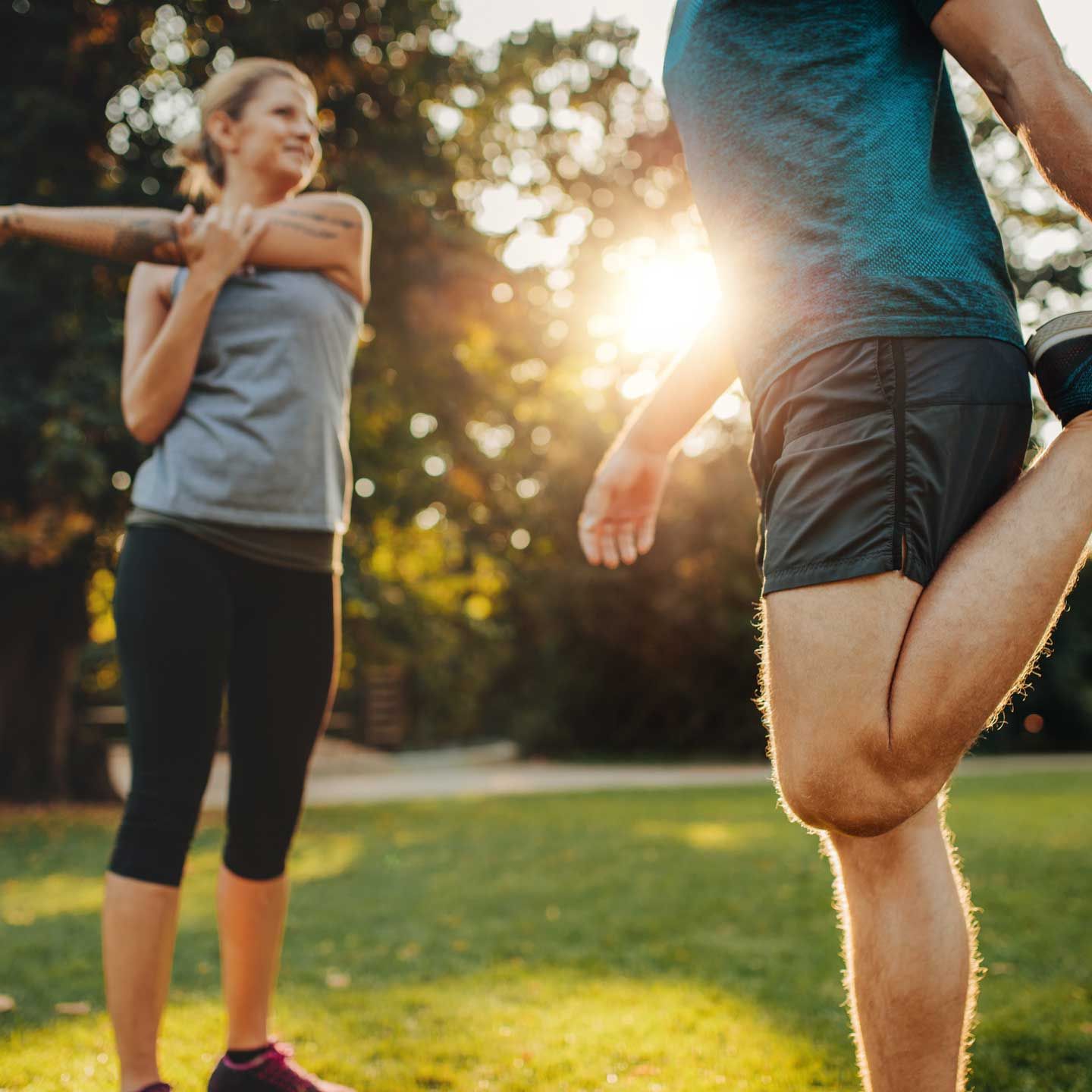 Two people stretching in a park: one with arm across chest, other holding leg up. Sunlight in background.