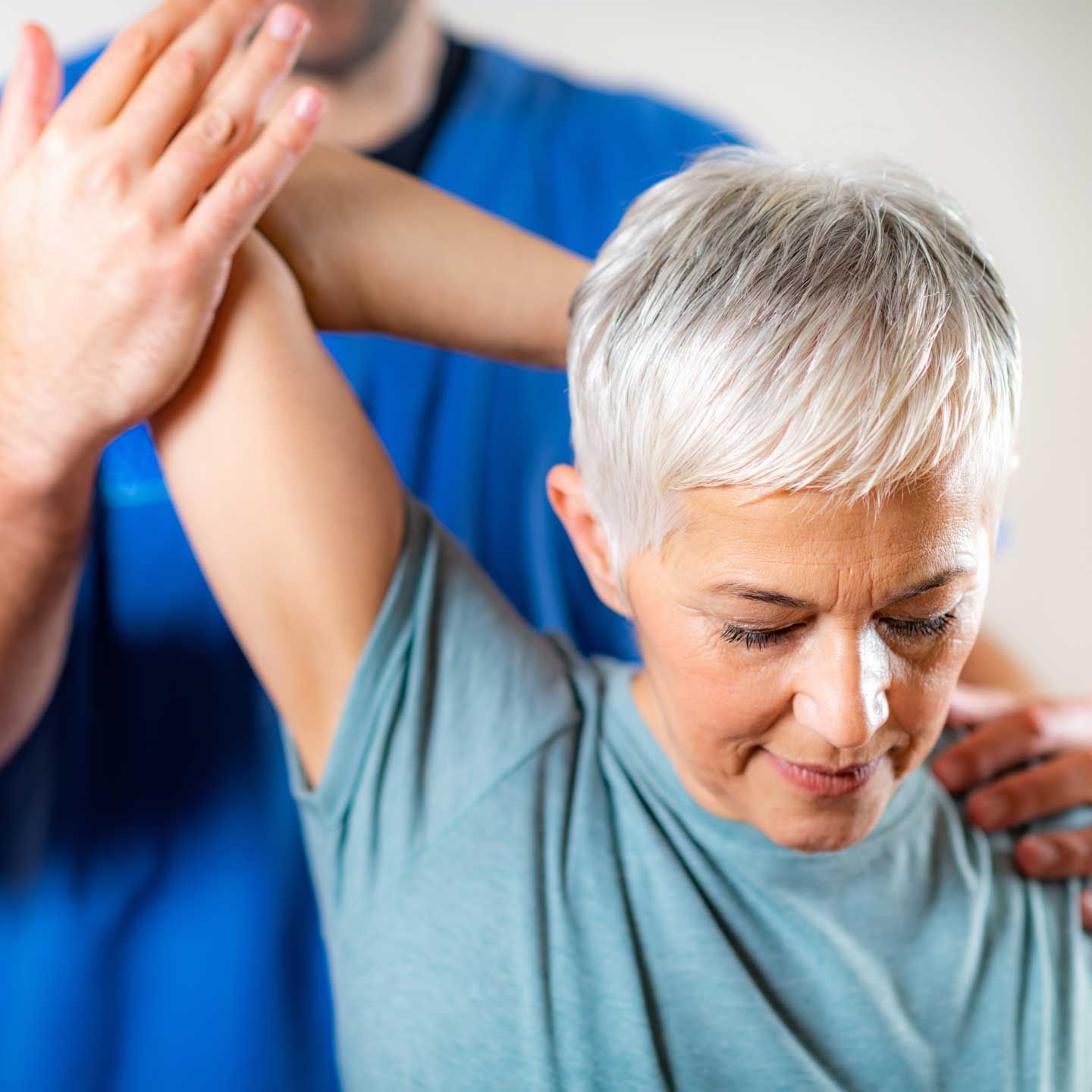Therapist assisting a person with raising arm; indoor, white background, blue and teal clothing.