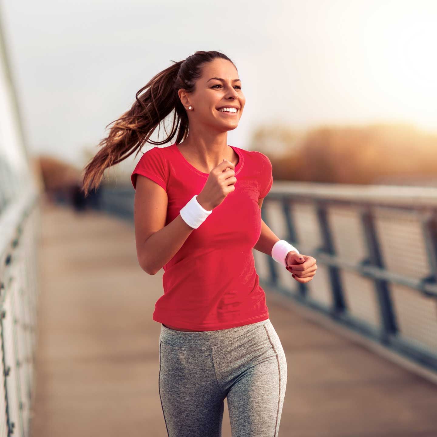 Woman running on a bridge, wearing red shirt and gray pants, smiling, sunlight.
