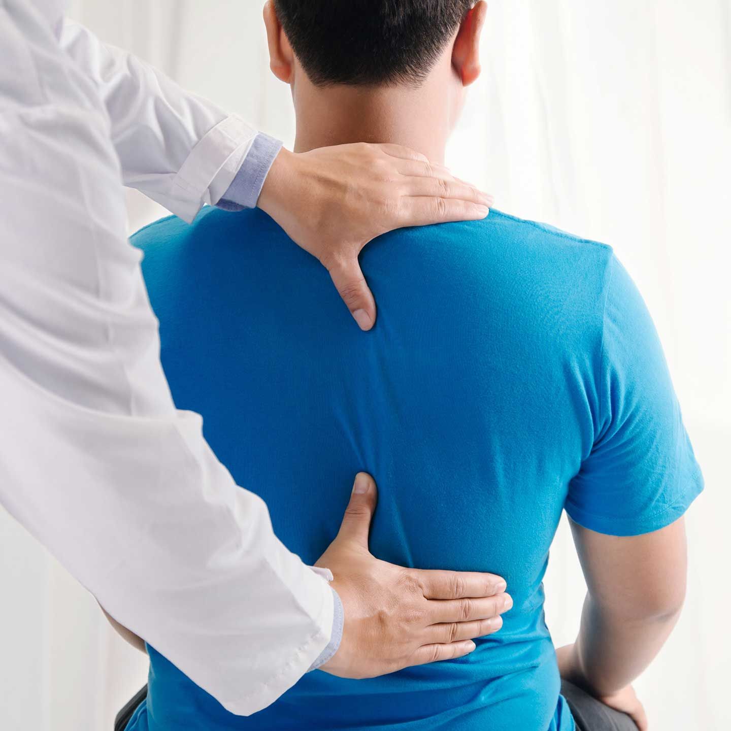Doctor examining a patient's back. Hands on the patient's upper back. Blue shirt, white coat. Indoor setting.