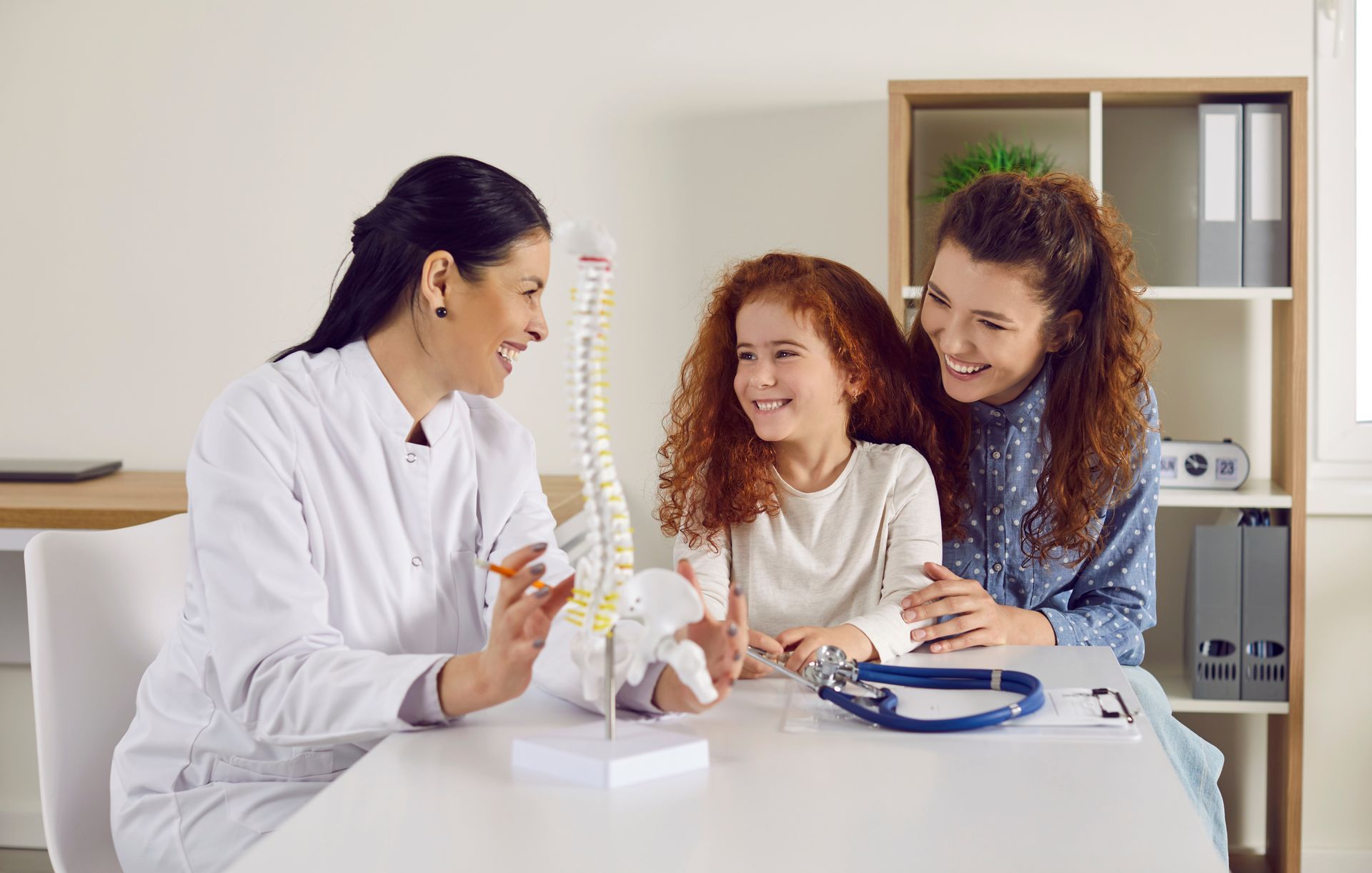 Doctor shows little girl anatomical model of spine standing on table in doctor's office.
