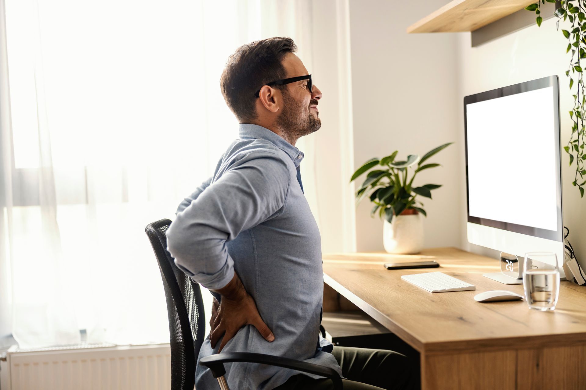 A man sitting at a home office desk, holding his lower back in discomfort.