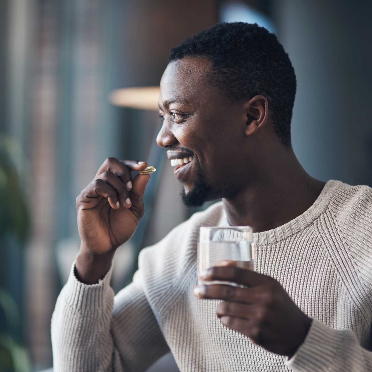 Man smiles while taking a pill with a glass of water indoors by a window.