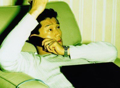 Man reclining in a green chair, talking on a telephone, looking up pensively.