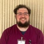 Smiling man with beard wearing maroon scrubs and glasses, ID badge visible.
