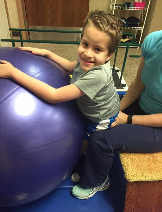 A smiling young boy with a braid is balancing on a therapy ball, being assisted by a seated person.