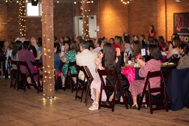 People seated at tables clapping in a room, likely an event. Wood floors, string lights, brick walls.