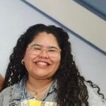 Woman with curly hair and glasses smiling, wearing a denim jacket.