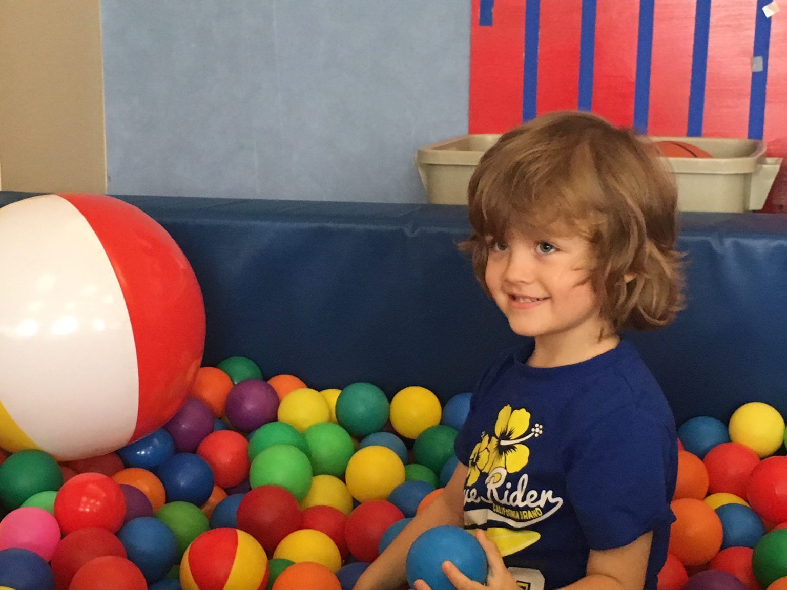 Young child smiling in a colorful ball pit, holding a blue ball near a beach ball.