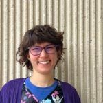 Woman with glasses smiles in front of a ribbed wall; wearing a purple cardigan, a blue shirt, and floral top.