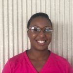 A smiling Black woman wearing glasses and a pink top, stands in front of a beige background.