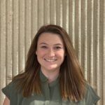 Woman with long brown hair smiles, wearing a green shirt, in front of a beige textured wall.