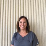 Woman with brown hair and gray scrubs smiles in front of a ribbed beige wall.