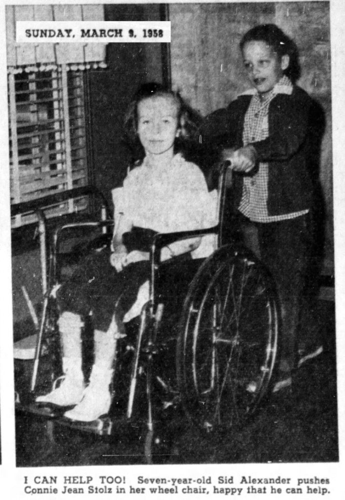 Connie Jean Steele in a wheelchair, pushed by a smiling kid, in a room. March 5, 1958.