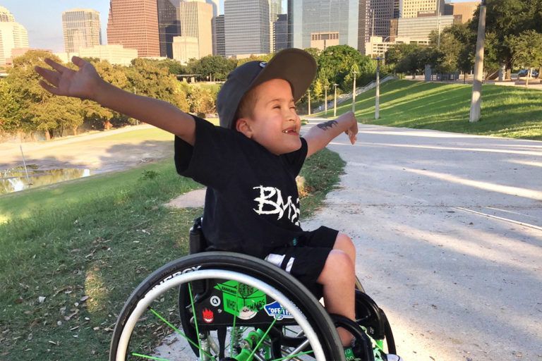 Boy in wheelchair with arms outstretched, smiling. He's in a park, downtown buildings in the background.